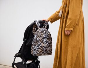 A woman in a yellow coat with their hand on a pushchair with a leopard print baby changing backpack hung on it