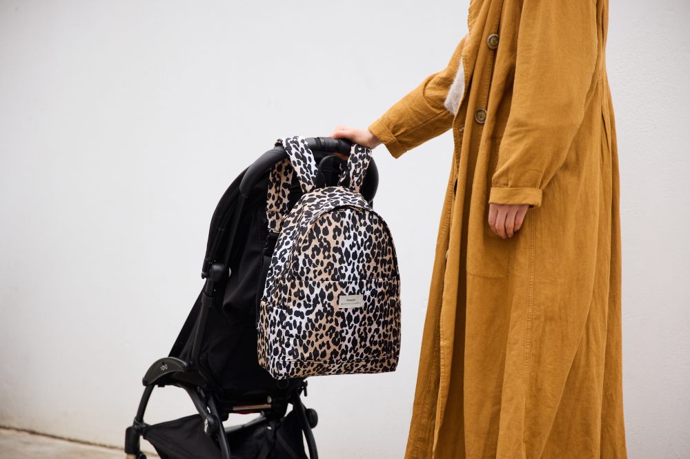 A woman in a yellow coat with their hand on a pushchair with a leopard print baby changing backpack hung on it