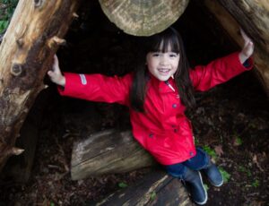A young girl sat in a tree trunk wearing a red rain coat and wellies