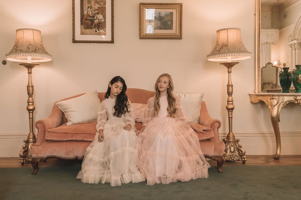 Two girls sat on a sofa wearing long dresses by Petite Amalie