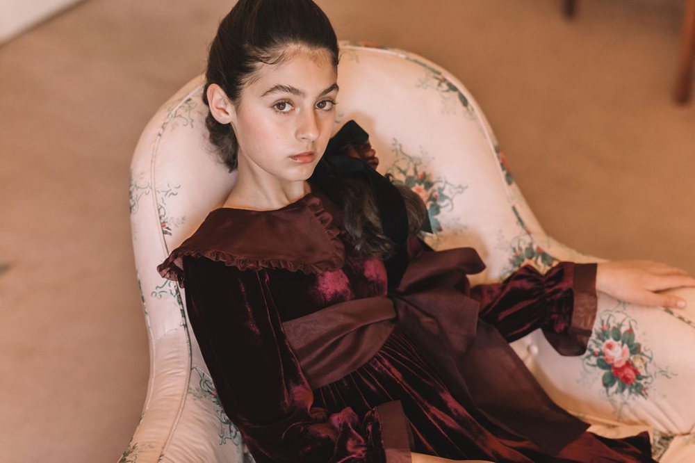 A girl sat in a chair wearing a long burgundy dress by Petite Amalie 