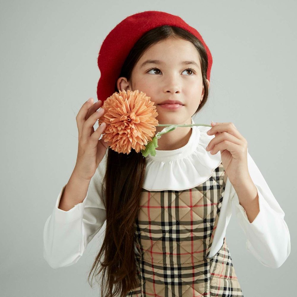 A young girl wearing a red beret holding up a pink flower 