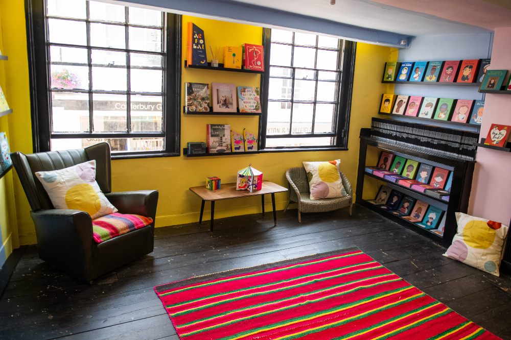 A room in a shop with yellow walls displaying books and a reading chair and table 