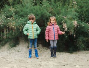 Two young children stood on a beach wearing wellies and coats by Muddy Puddles