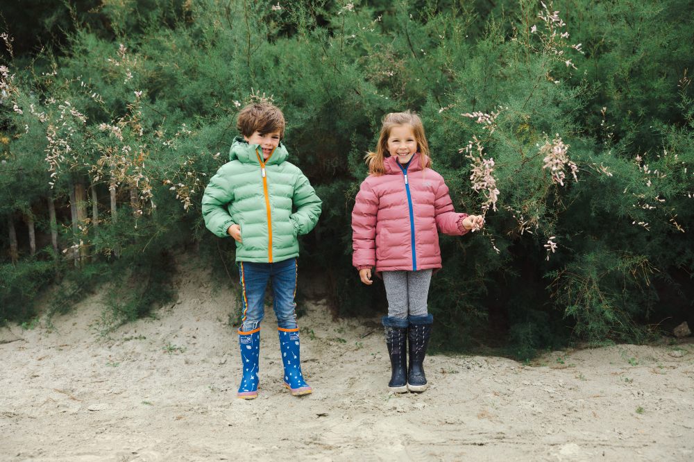 Two young children stood on a beach wearing wellies and coats by Muddy Puddles