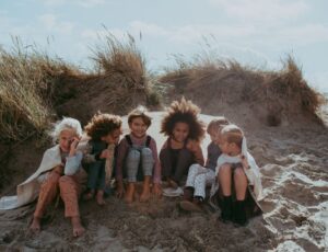 A group of young children sitting ion a sand dune under a blanket wearing outfits by Turtledove London