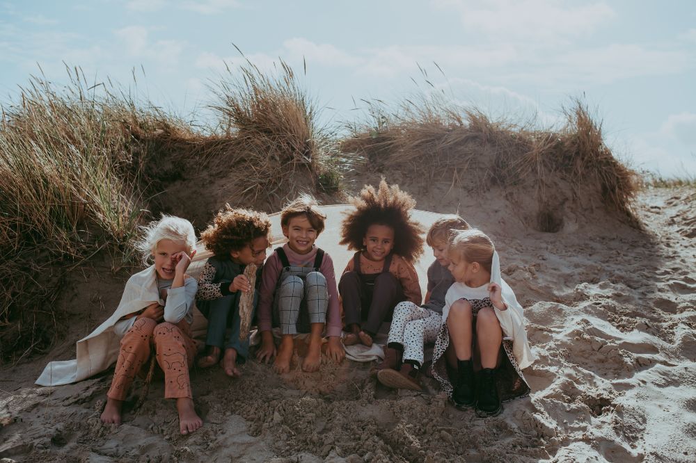 A group of young children sitting ion a sand dune under a blanket wearing outfits by Turtledove London