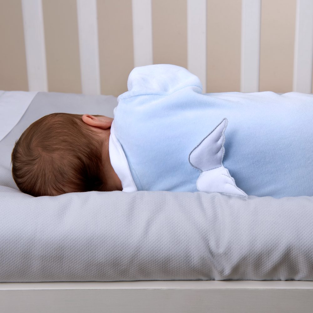 A baby lying in a cot wearing a blue babygro with wings on the back