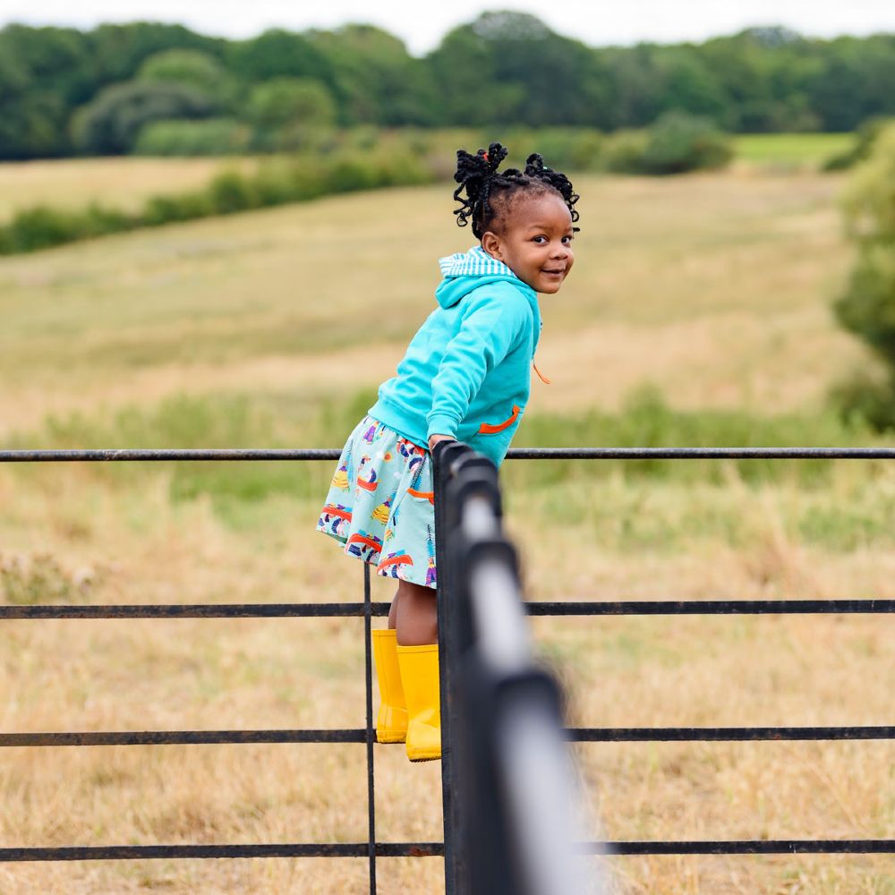 A young girl stood on a gate in a field wearing a turquoise top, skirt and yellow wellington boots