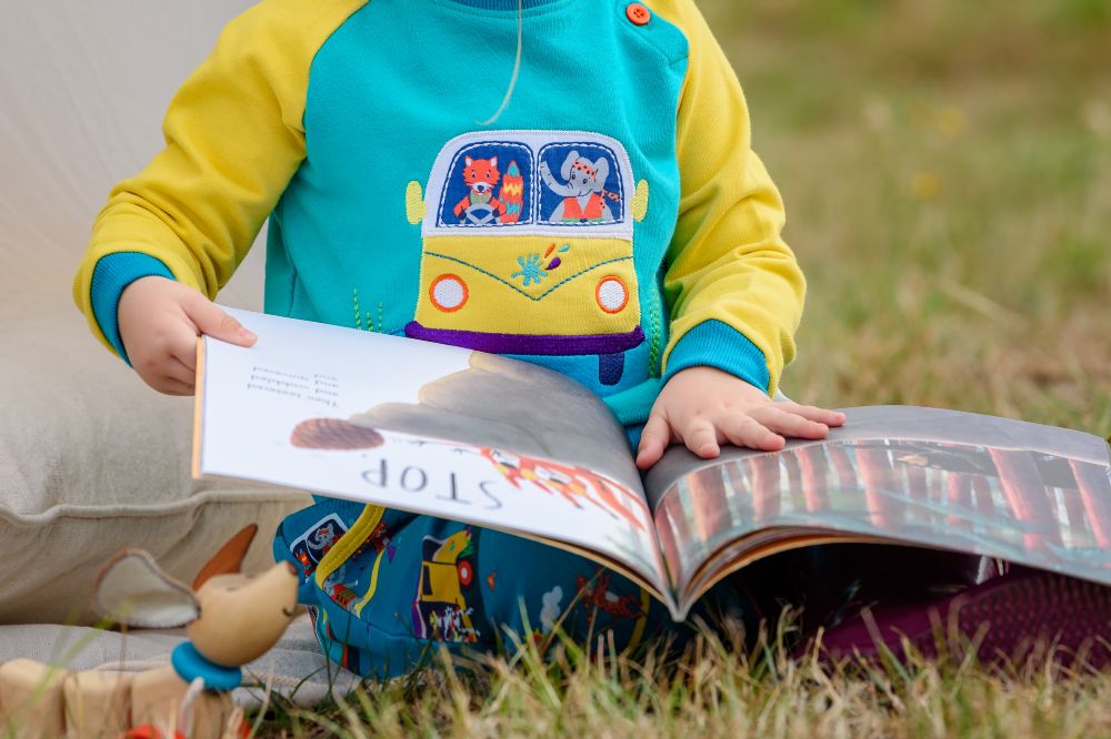 A child sat on grass reading a book and wearing a top with a campervan on the front by Ducky Zebra 