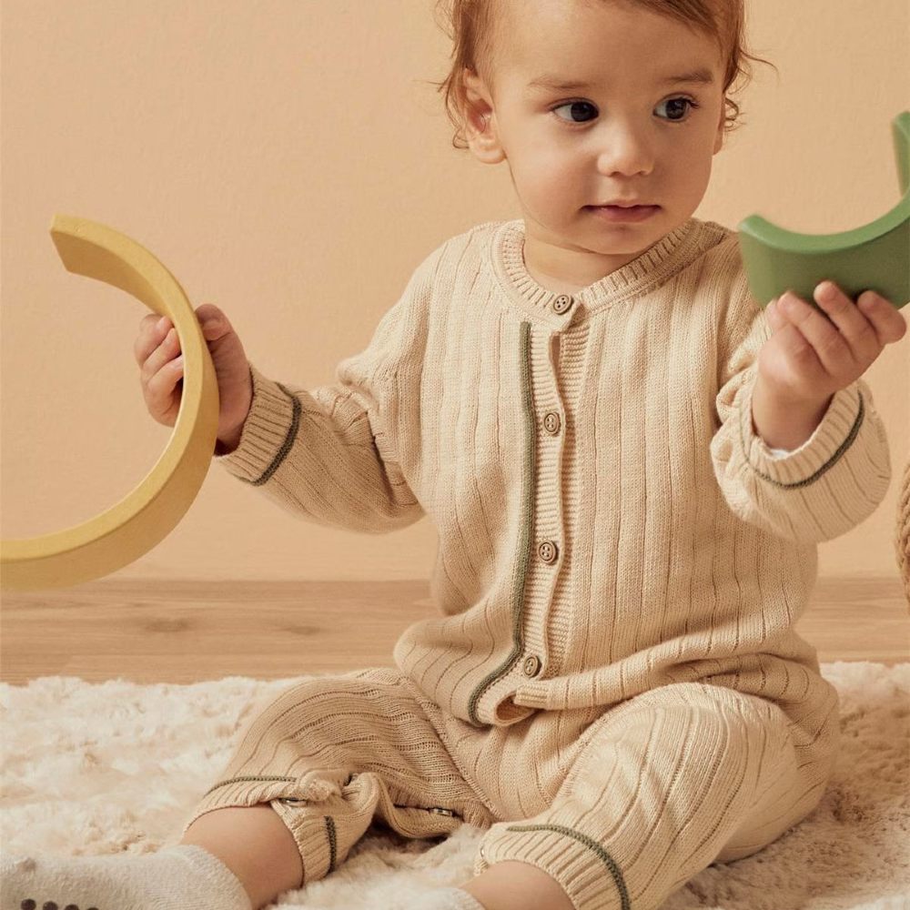 A baby sat on the floor holding toys wearing a beige bodysuit