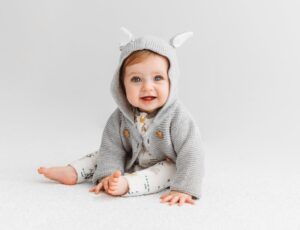 A baby sat on the floor wearing a grey hooded cardigan and white trousers by Kit & Kin