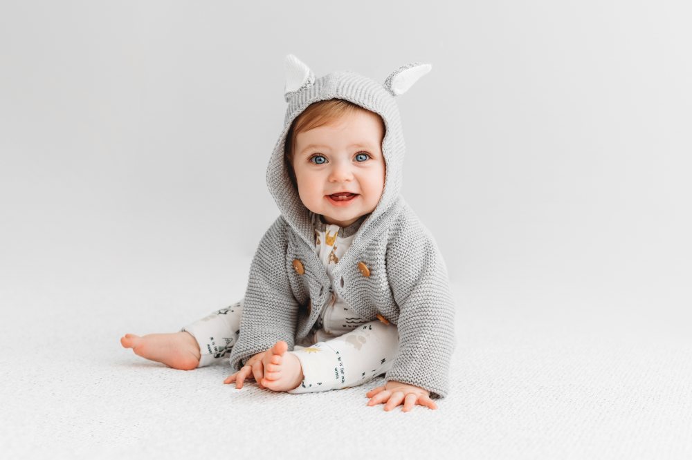 A baby sat on the floor wearing a grey hooded cardigan and white trousers by Kit & Kin