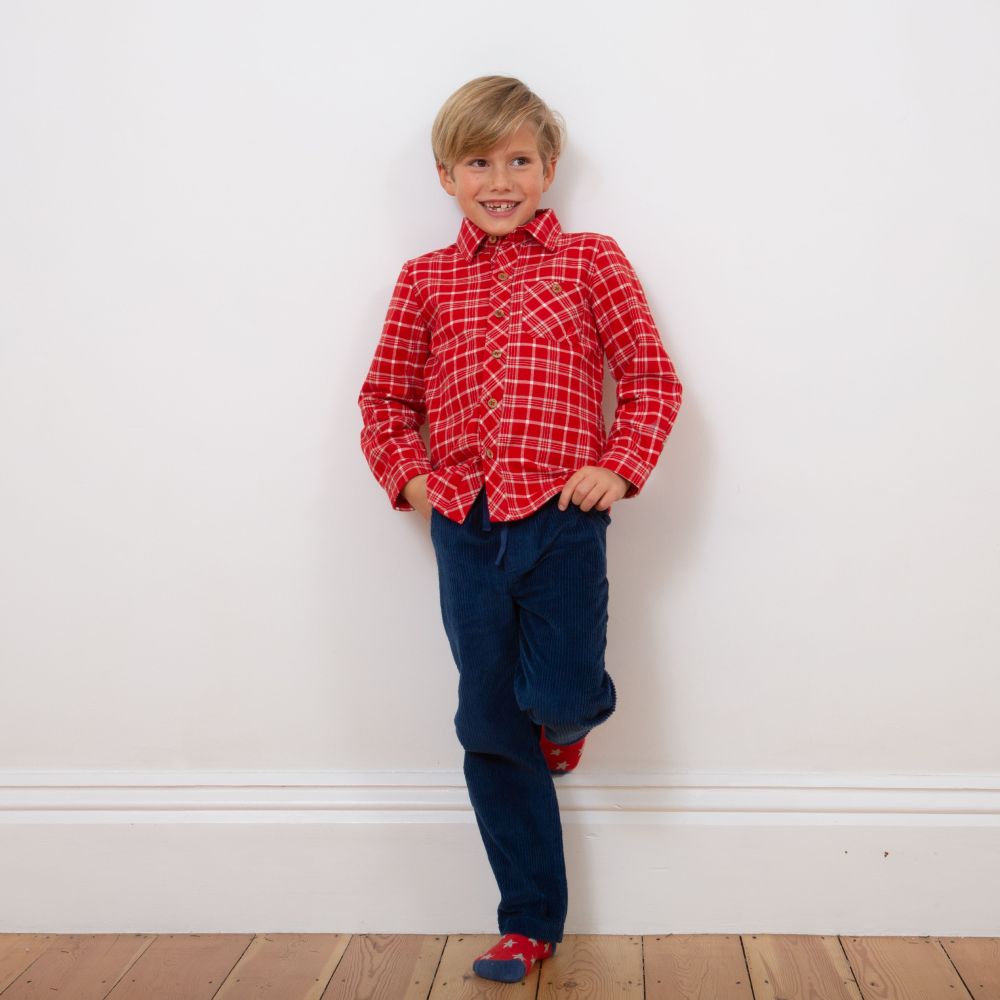 A young boy stood against a white wall wearing a read checked shirt and navy trousers 