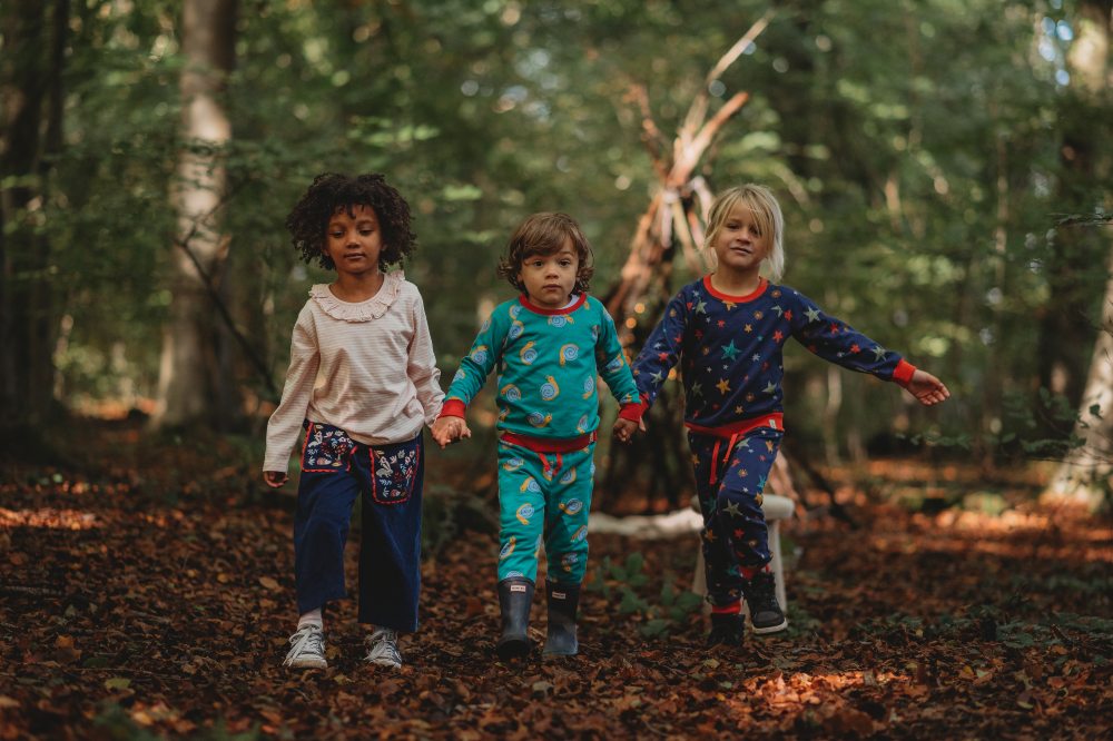 Three children holding hands walking through a wood wearing outfits by Lilly + Sid