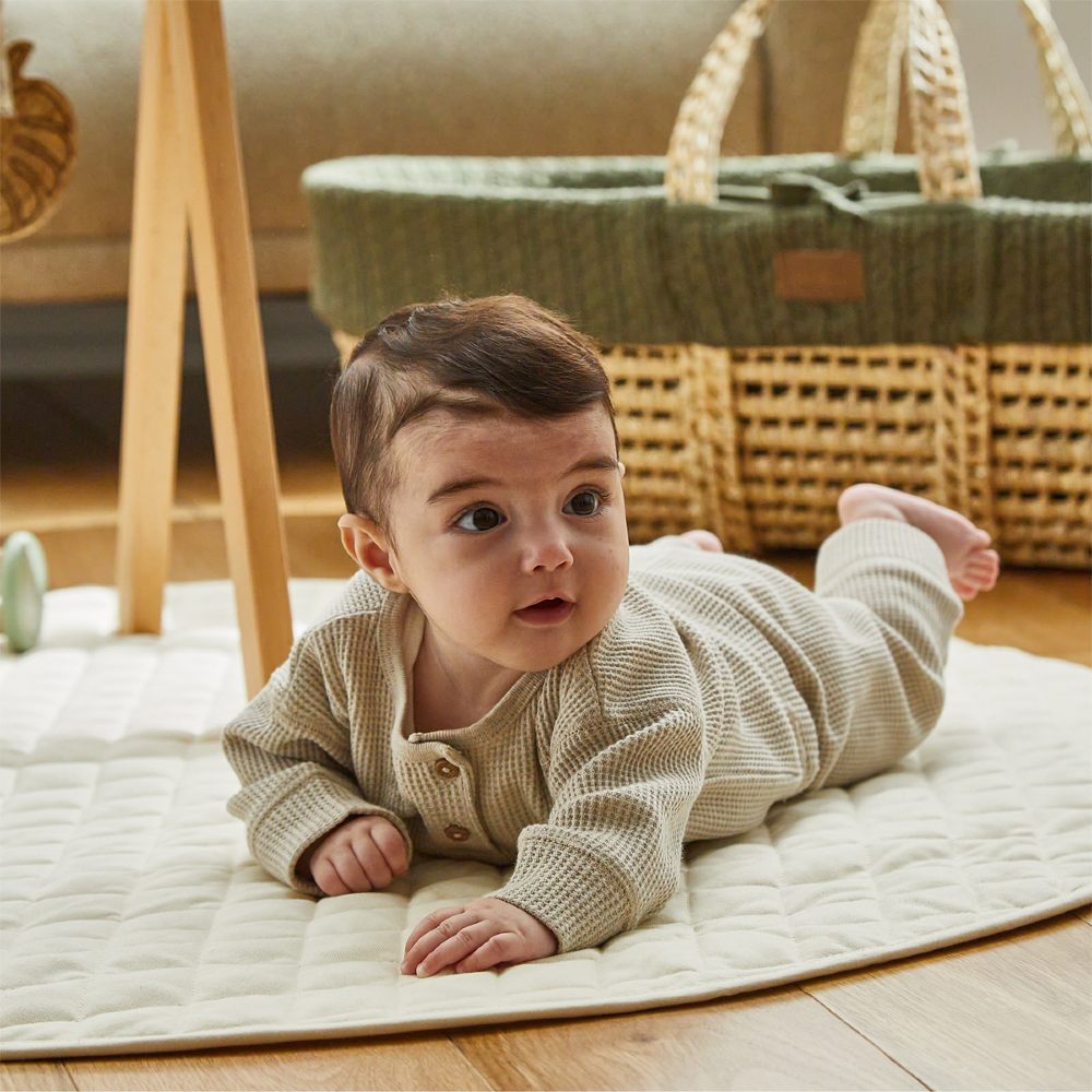 A young baby lying on a rug beside a Moses Basket by The Little Green Sheep