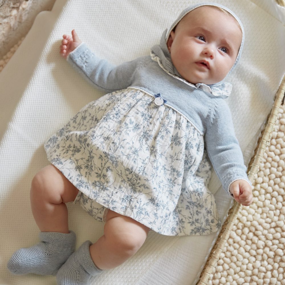 A baby lying on its back wearing a patterned dress and blue cardigan 