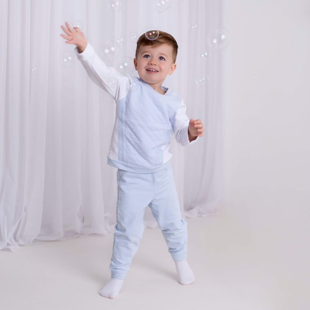 A young boy raising his arm to catch bubbles wearing a pale blue top and matching trousers 