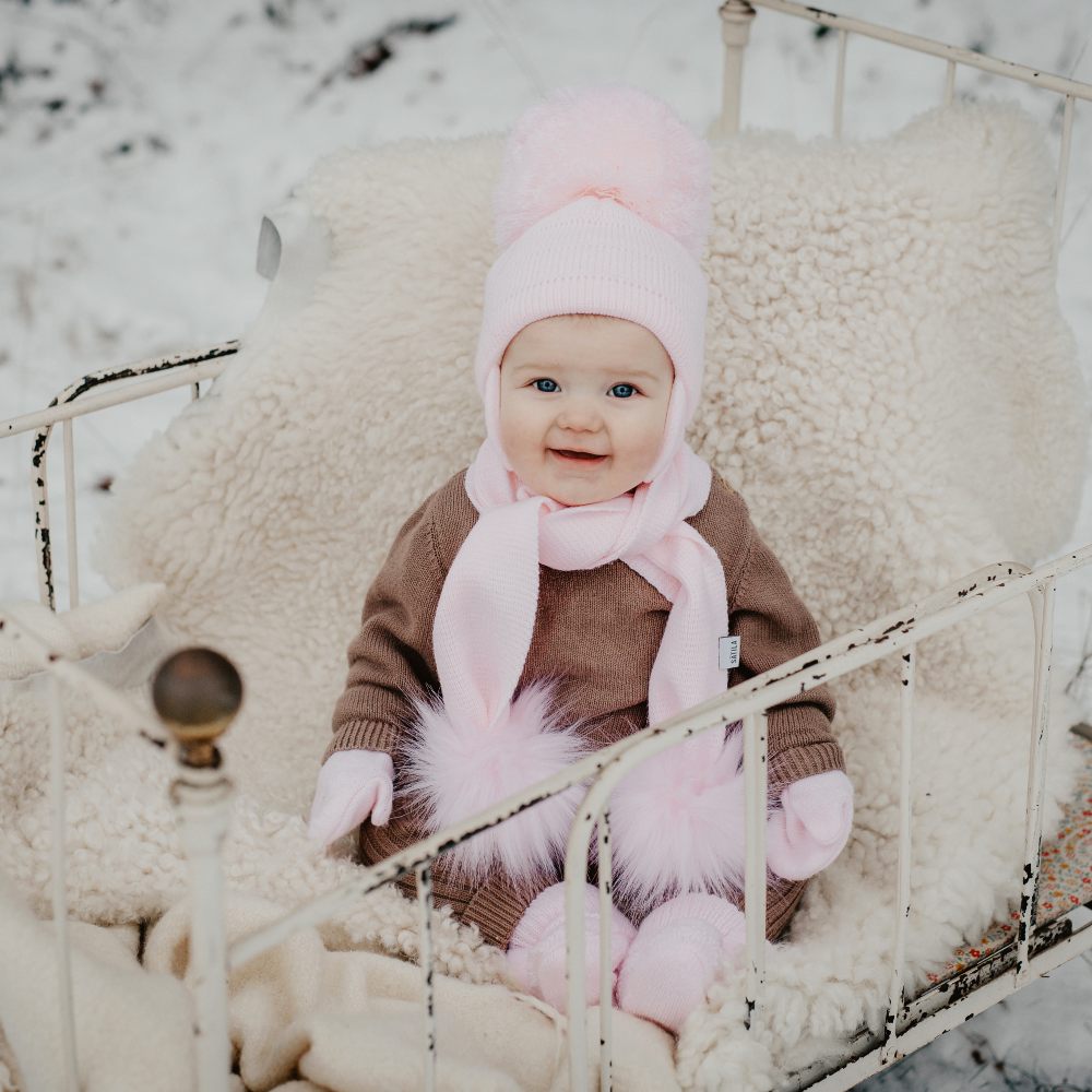 A baby sat in a snowy scene wearing a hat and scarf with fur pompoms