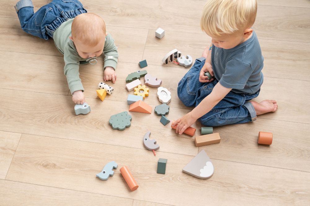 A young child and a baby on the floor playing with wooden toys from the Happy Home 24 Collection by Done by Deer