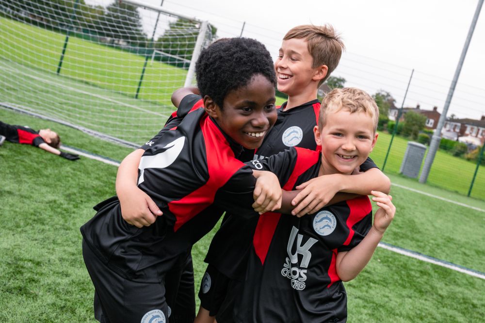 A group of boys in black and red sports kit hugging on a football pitch 