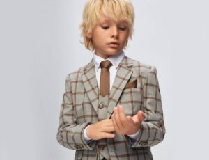 A young boy with blonde hair wearing a three-piece suit and tie and adjusting his sleeve