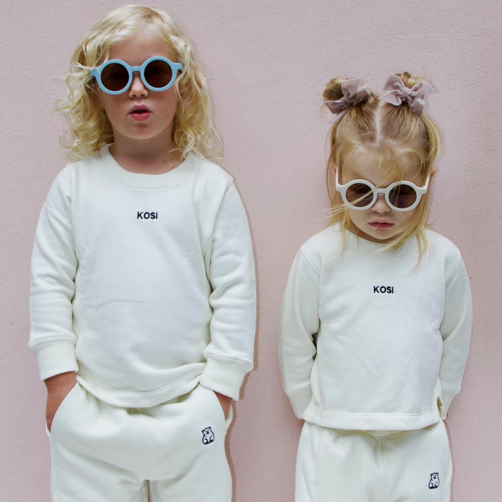 Two children stood against a pink wall wearing sunglasses and white tops and trousers with Kosi Kids branding 