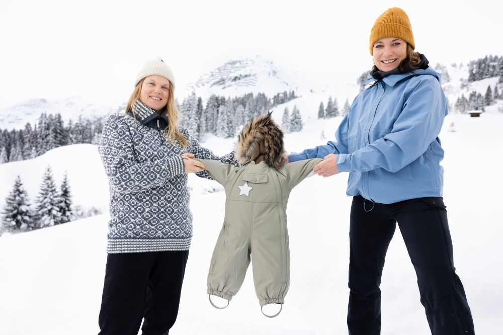 Two women stood outside in the snow holding up a Mini A Ture snowsuit