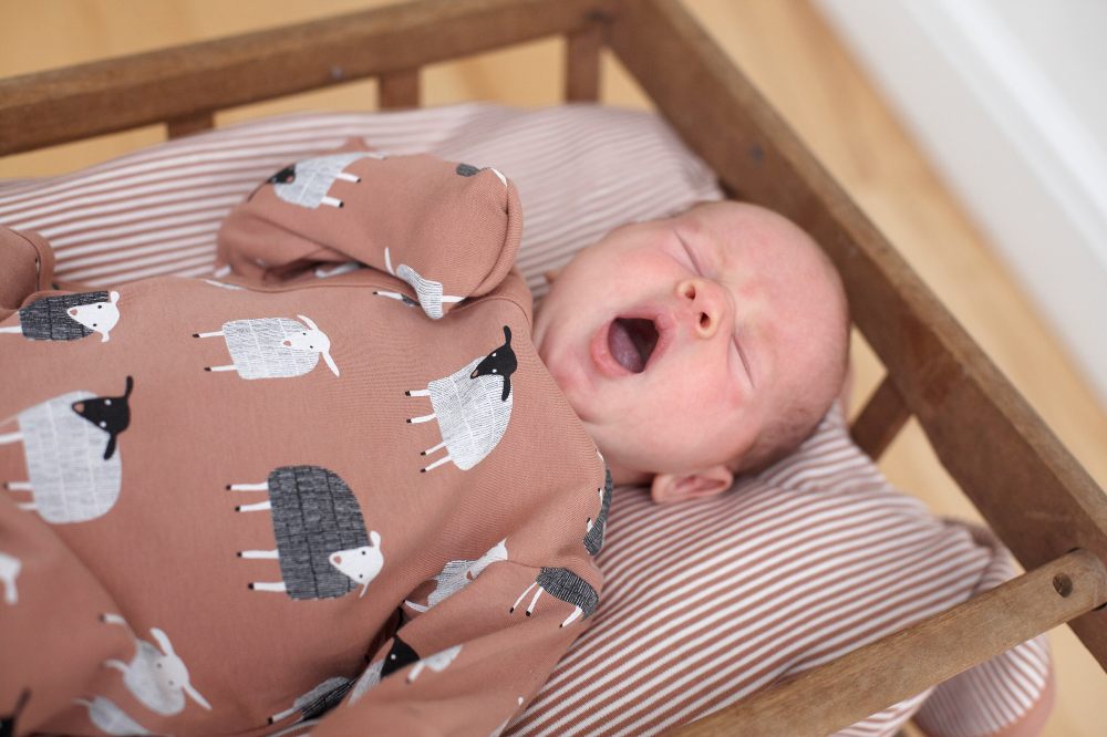 A baby lying in a crib yawning wearing a babygro with sheep on 