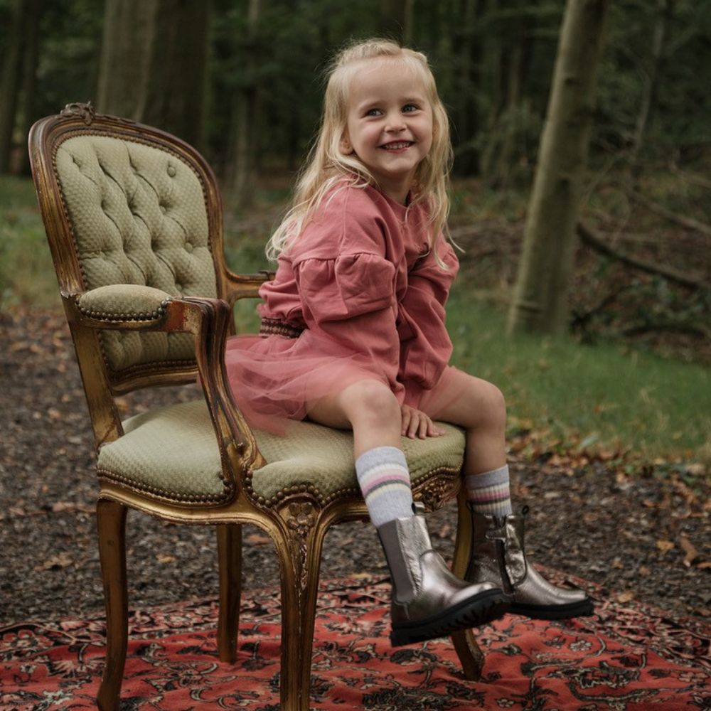 A young girl in a pink dress and metallic boots sat outside on a chair in a wood 