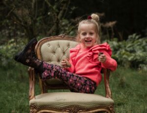A girl sat in a wood on an ornate chair with her legs on the chair's arm