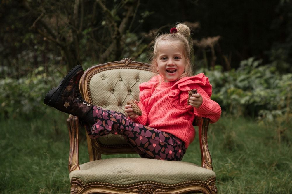 A girl sat in a wood on an ornate chair with her legs on the chair's arm
