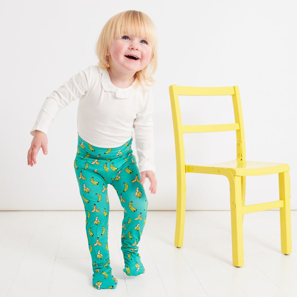 A young child in a white top and turquoise banana print leggings stood beside a yellow chair 