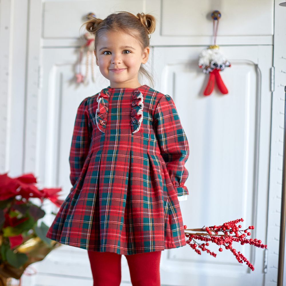 A young girl stood in a room decorated for Christmas wearing a red and green checked dress and red tights 