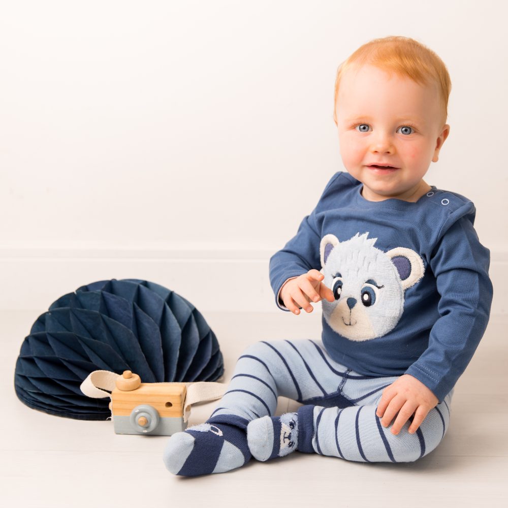 A baby sat on the floor wearing a blue top with a bear on and blue striped leggings 