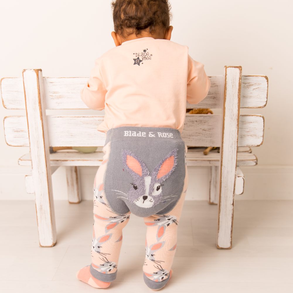 A baby stood up holding on to a white chair wearing a pink top and leggings with a bunny on the bottom 