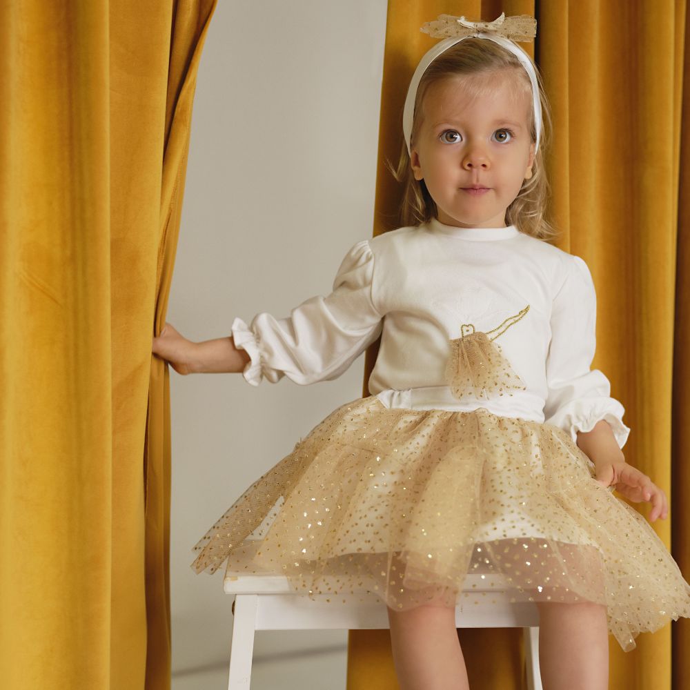 A young girl sat on a stool wearing a white and gold dress and holding back a yellow curtain
