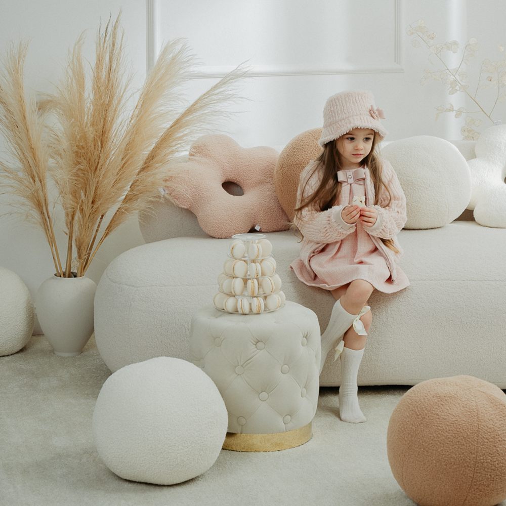 A young girl sat on a white sofa wearing a pink hat and dress and white knee socks 