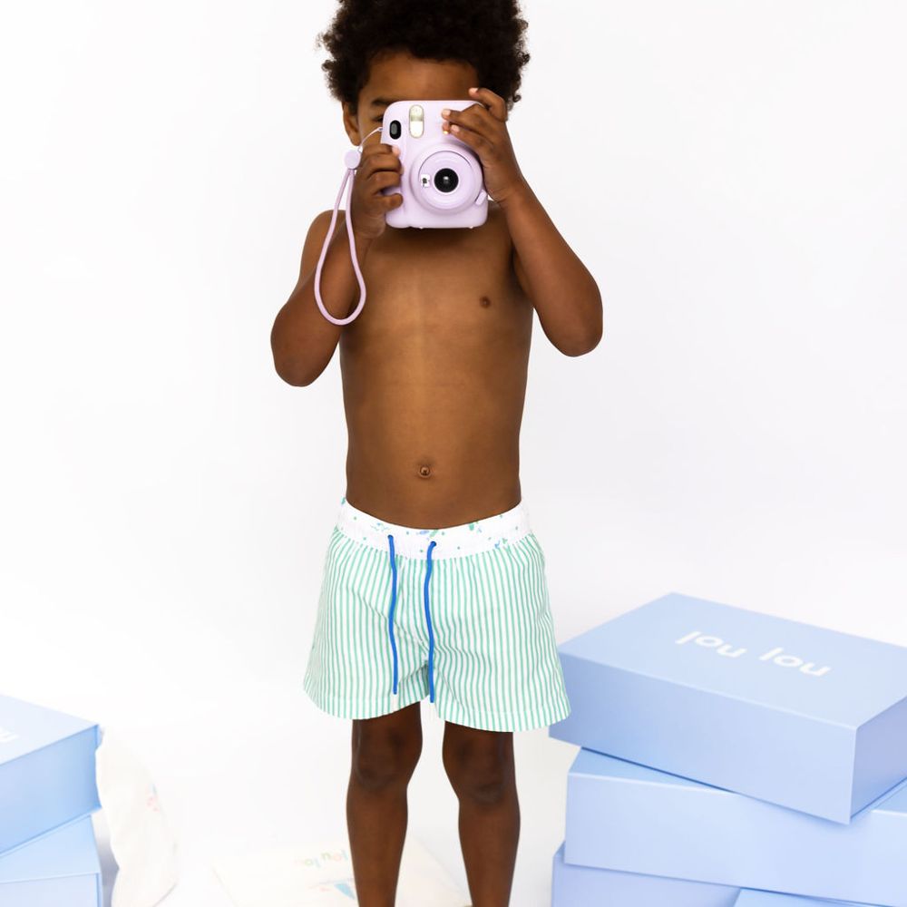 A young boy wearing blue striped swim shorts and holding a camera to his face