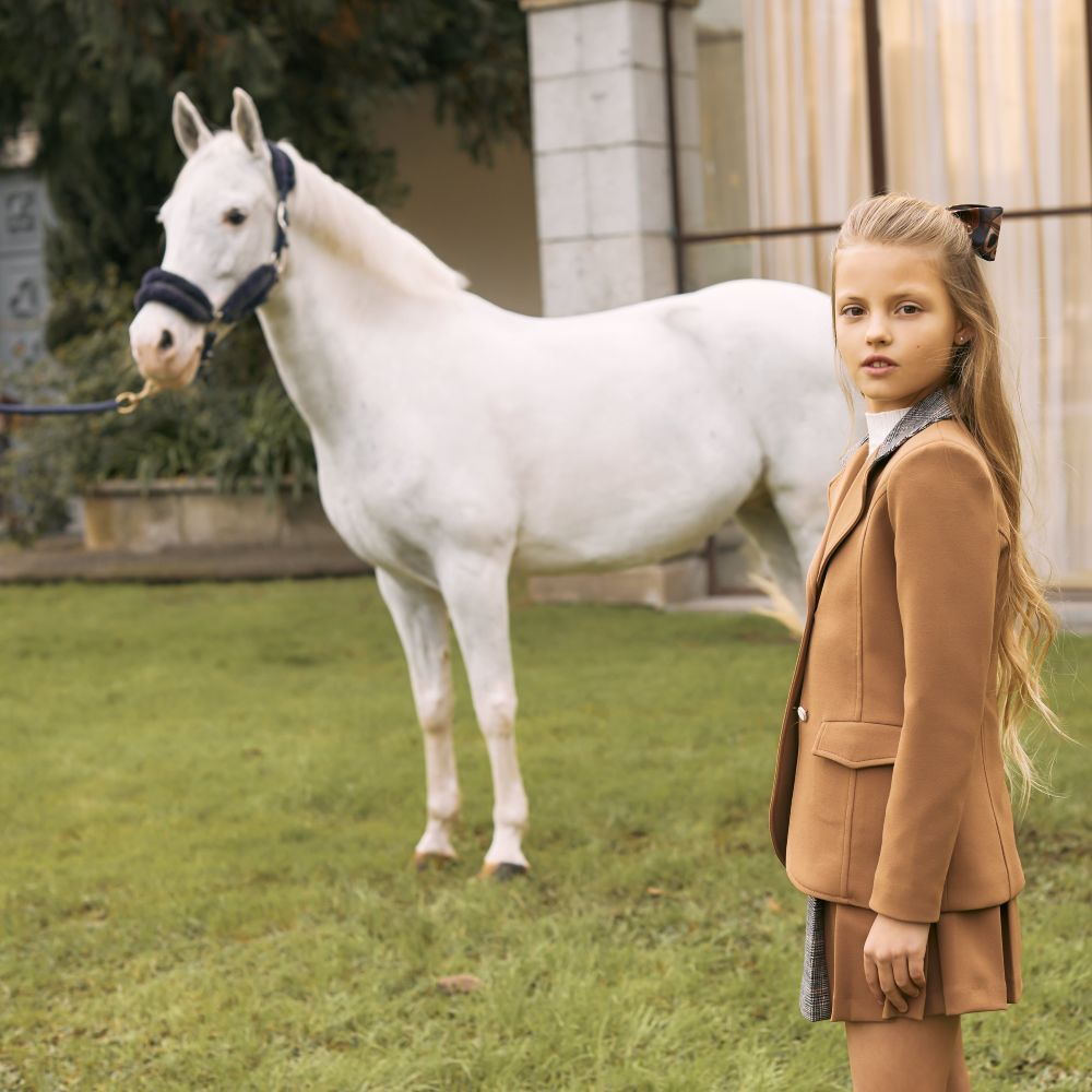 A girl wearing a beige jacket and skirt stood outside beside a white horse 