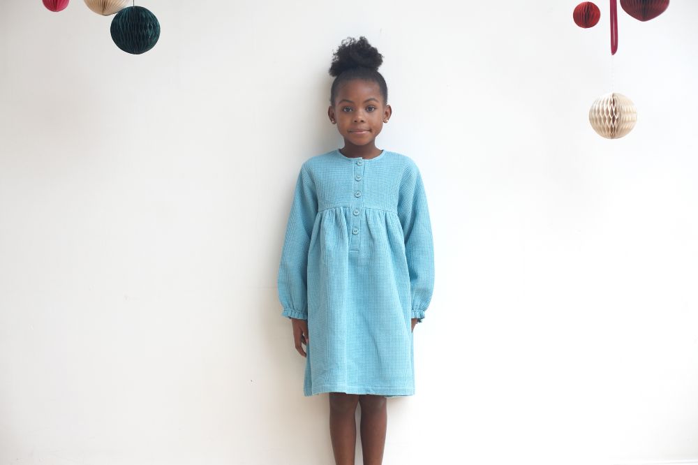 A young girl stood against a white wall wearing a pale blue dress 