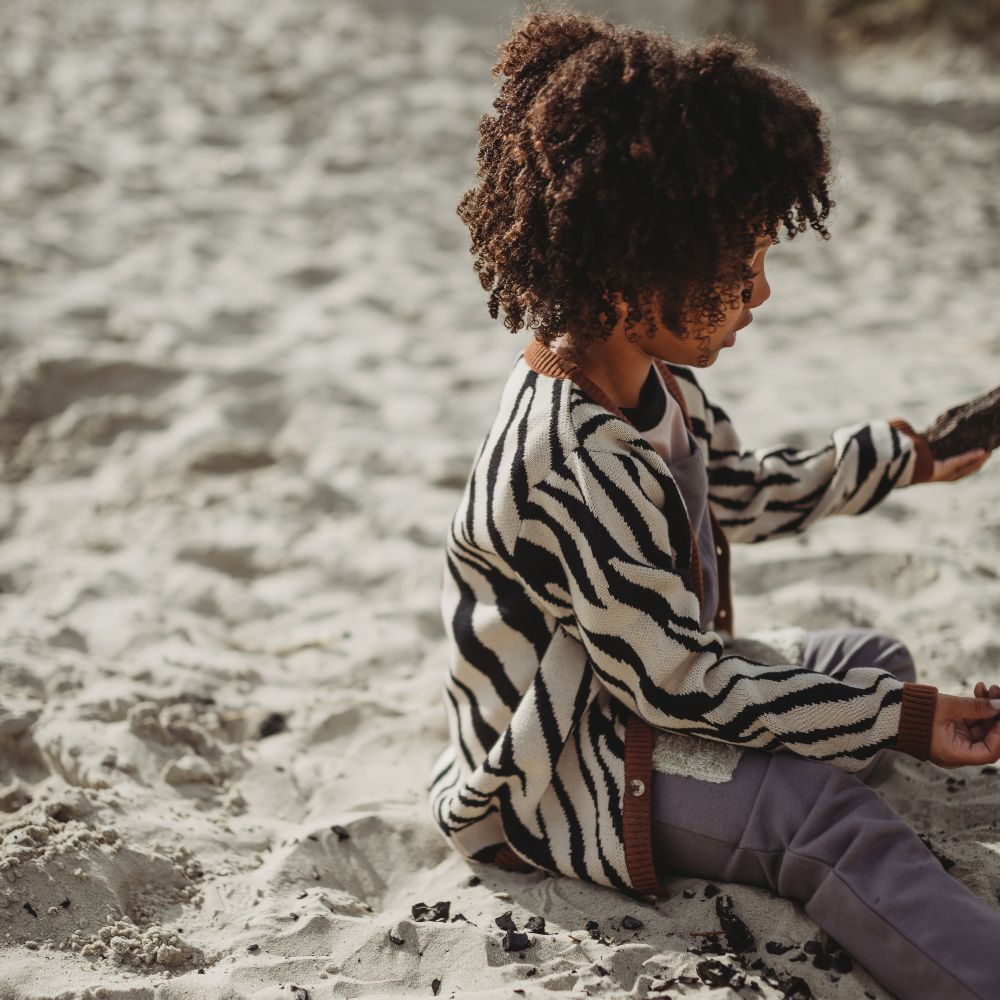 A young child sand on a beach wearing a zebra print cardigan and grey trousers 