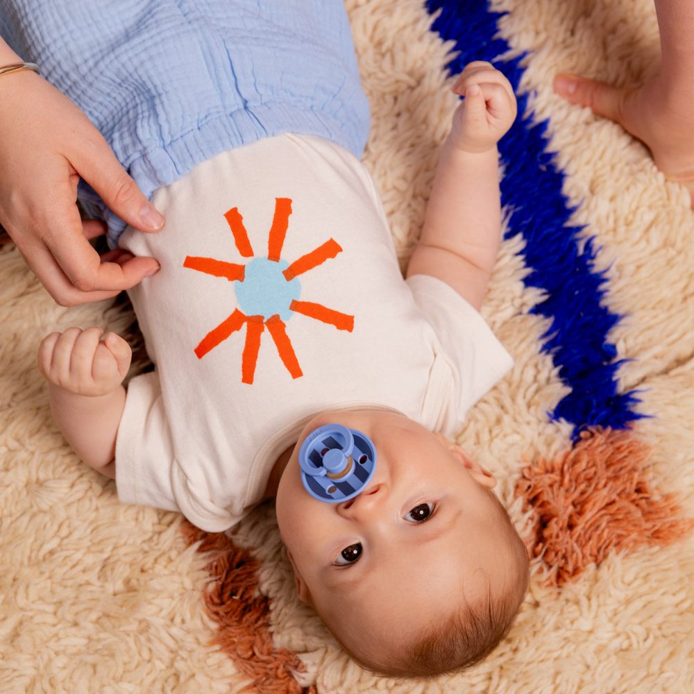 A baby lying on a rug with a blue striped pacifier in their mouth 