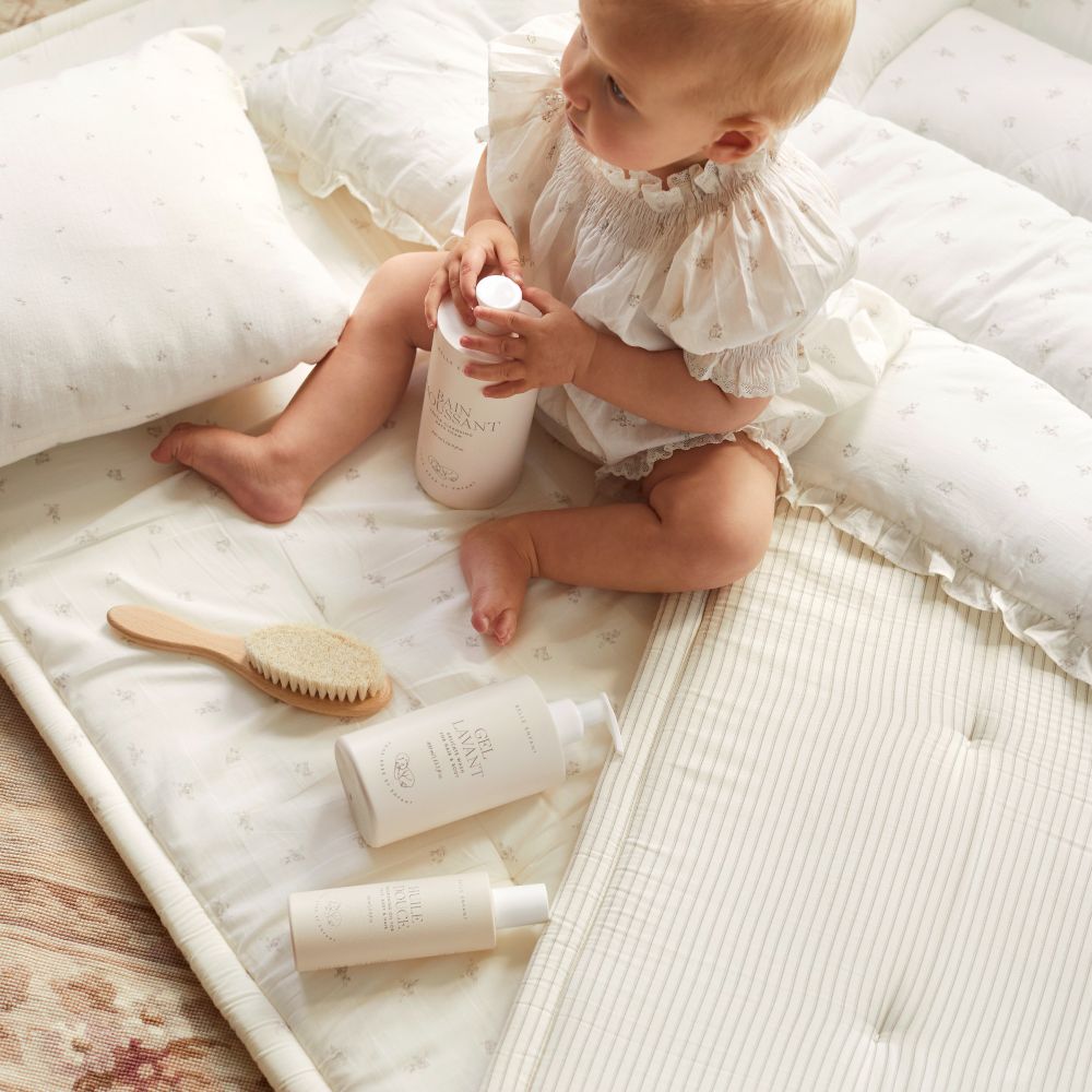 A baby sat on bedding holding a bottle 