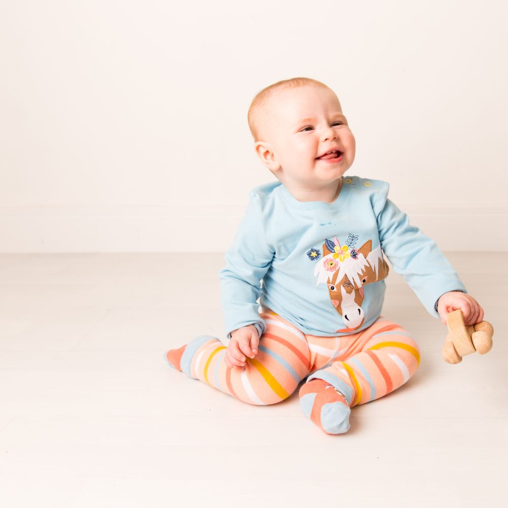 A baby sat on the floor wearing striped leggings and a blue top with a horse motif on the front