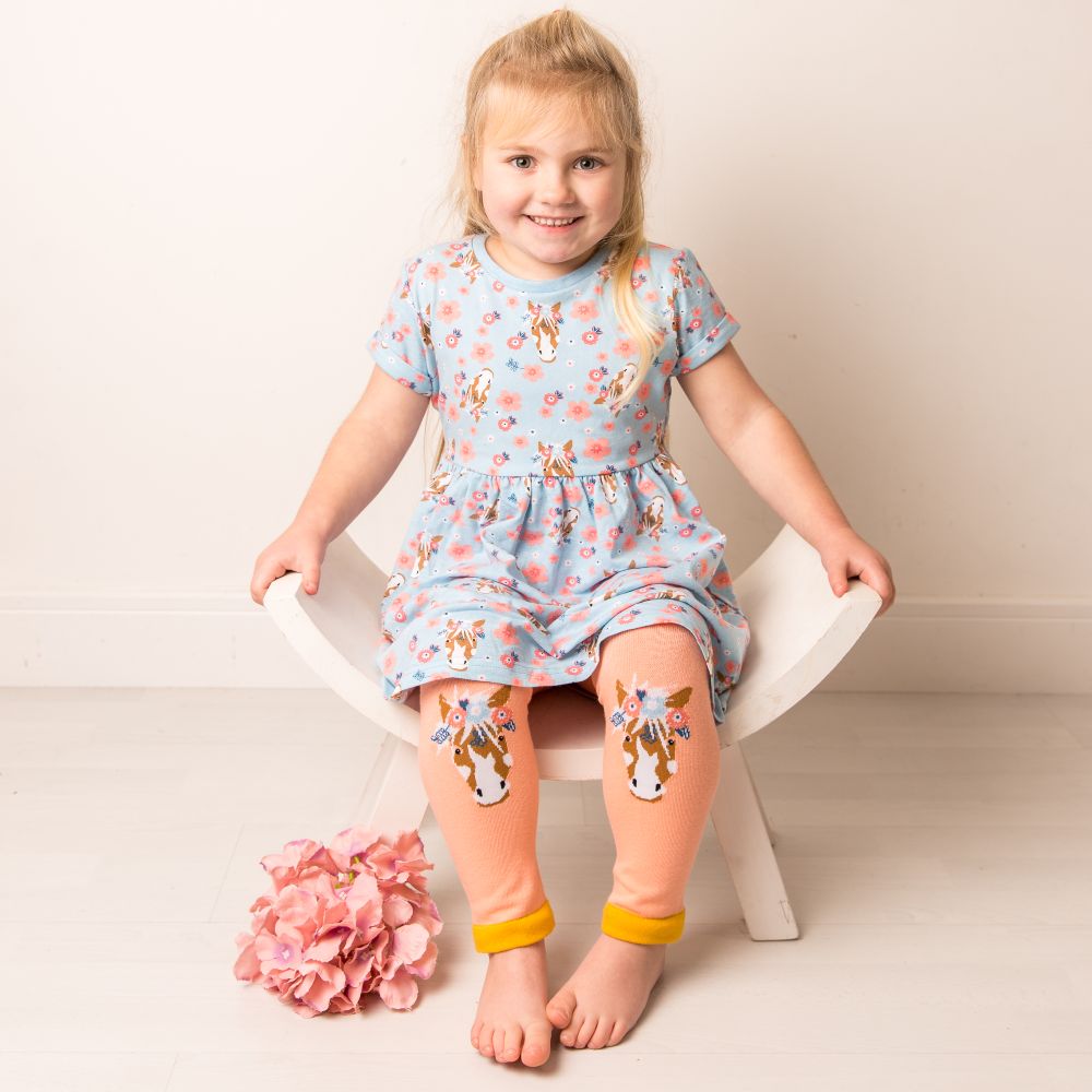 A girl sat on a chair wearing a dress and leggings with horse motifs on the knees