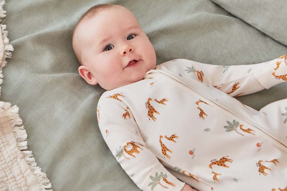 A baby lying down wearing a giraffe print babygro by MORI