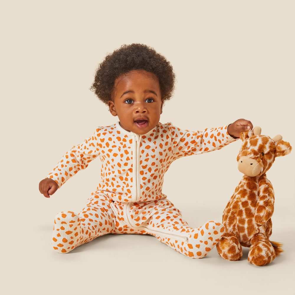 A young baby sat on the floor wearing a giraffe print babygro and reaching out for a giraffe soft toy