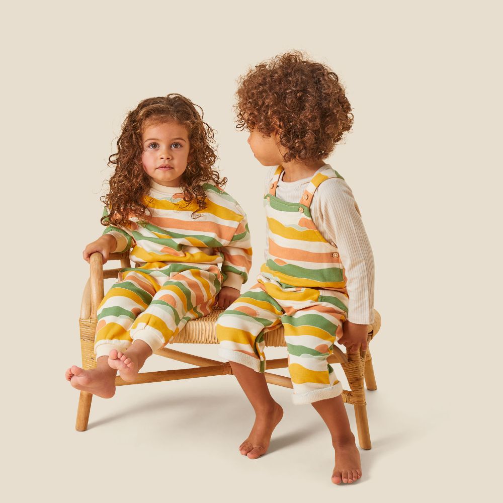 Two children sat on a wooden chair wearing orange, green and white patterned outfits