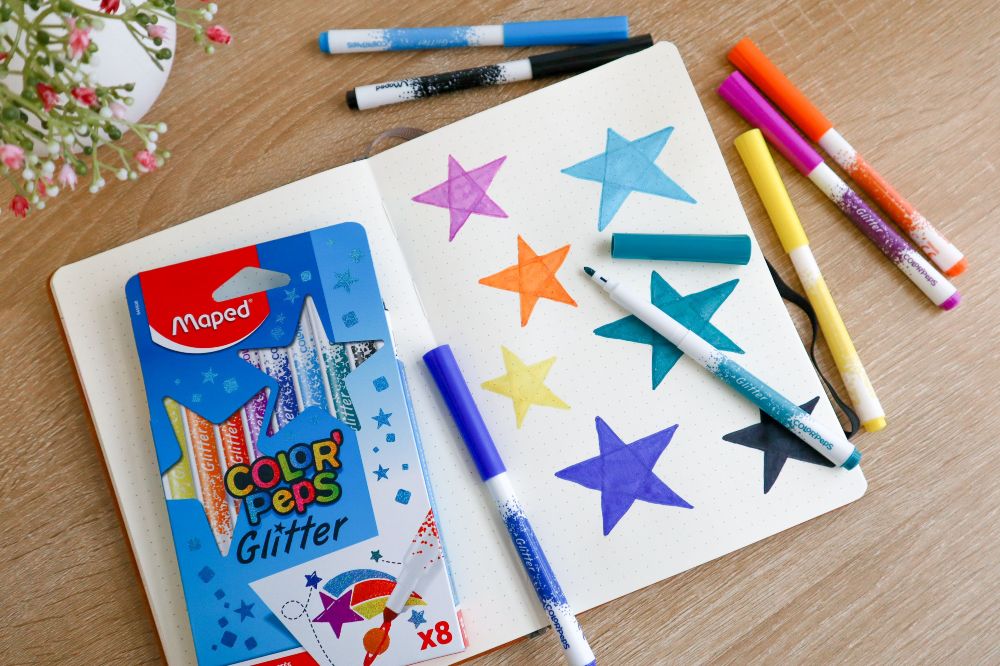 A notebook with a child's drawing beside a box of glitter colouring pens 